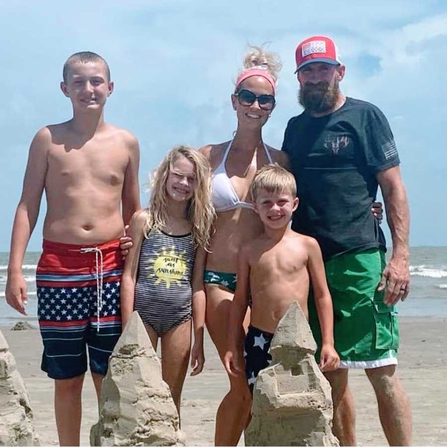 A woman stands at the beach with her family behind a sandcastle