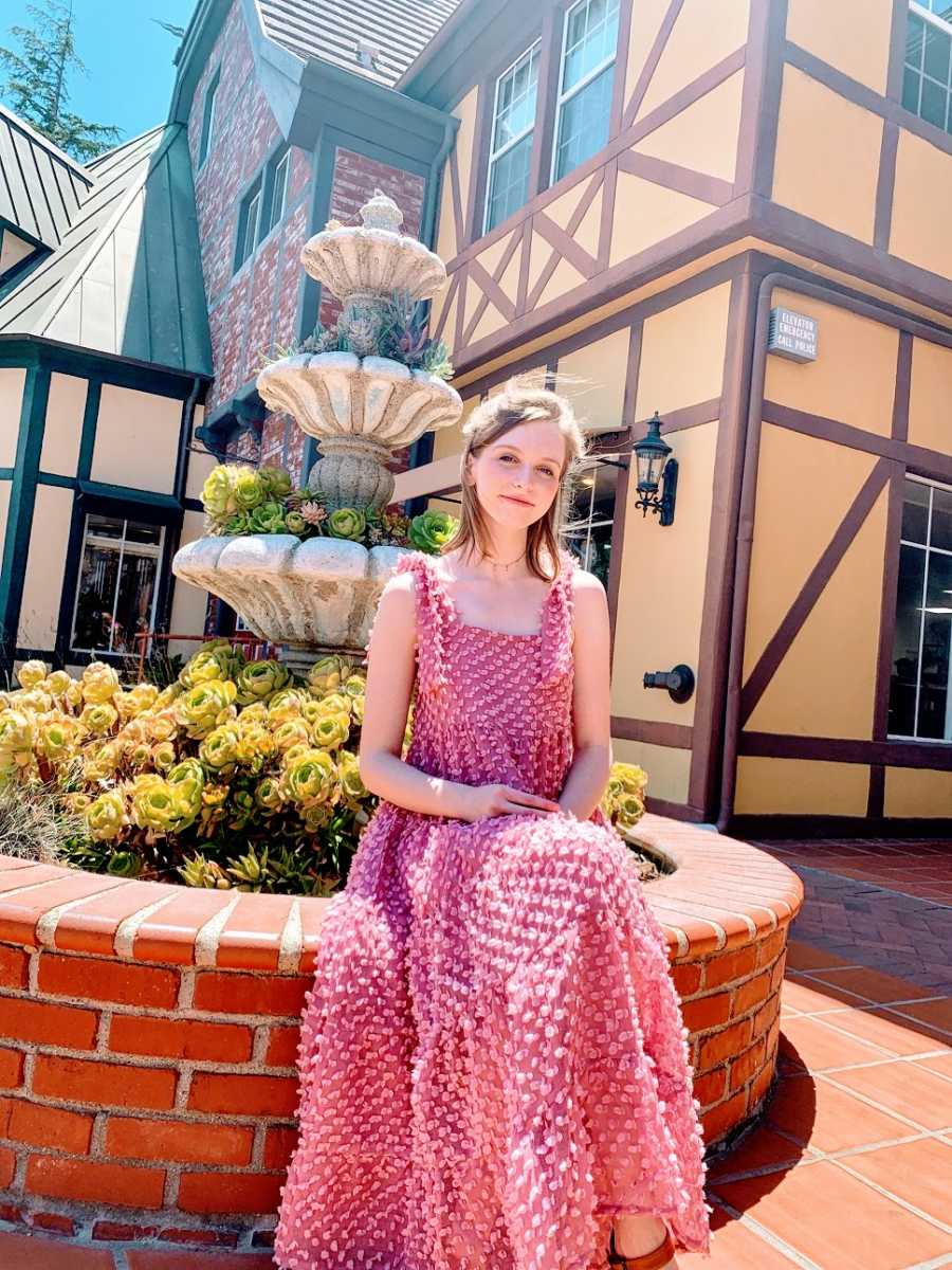 A young woman in a pink dress sits on a fountain