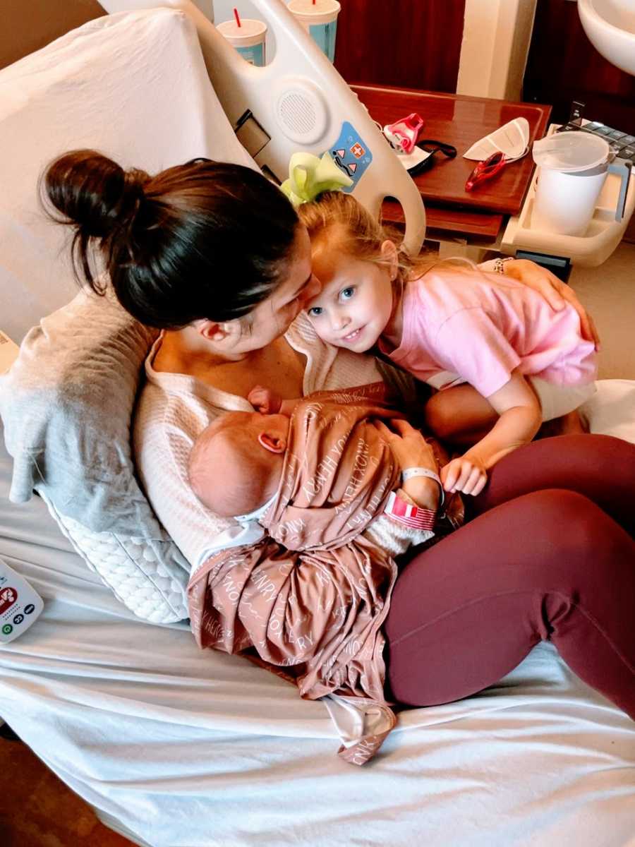 A mother sits in a hospital bed with her newborn and her daughter