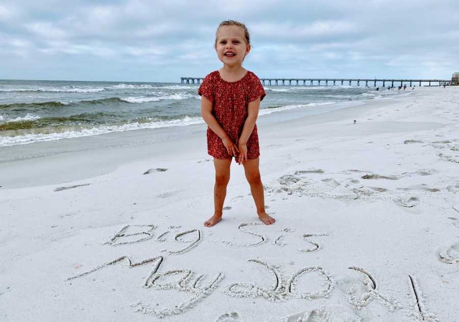A little girl stands on a beach with writing in the sand