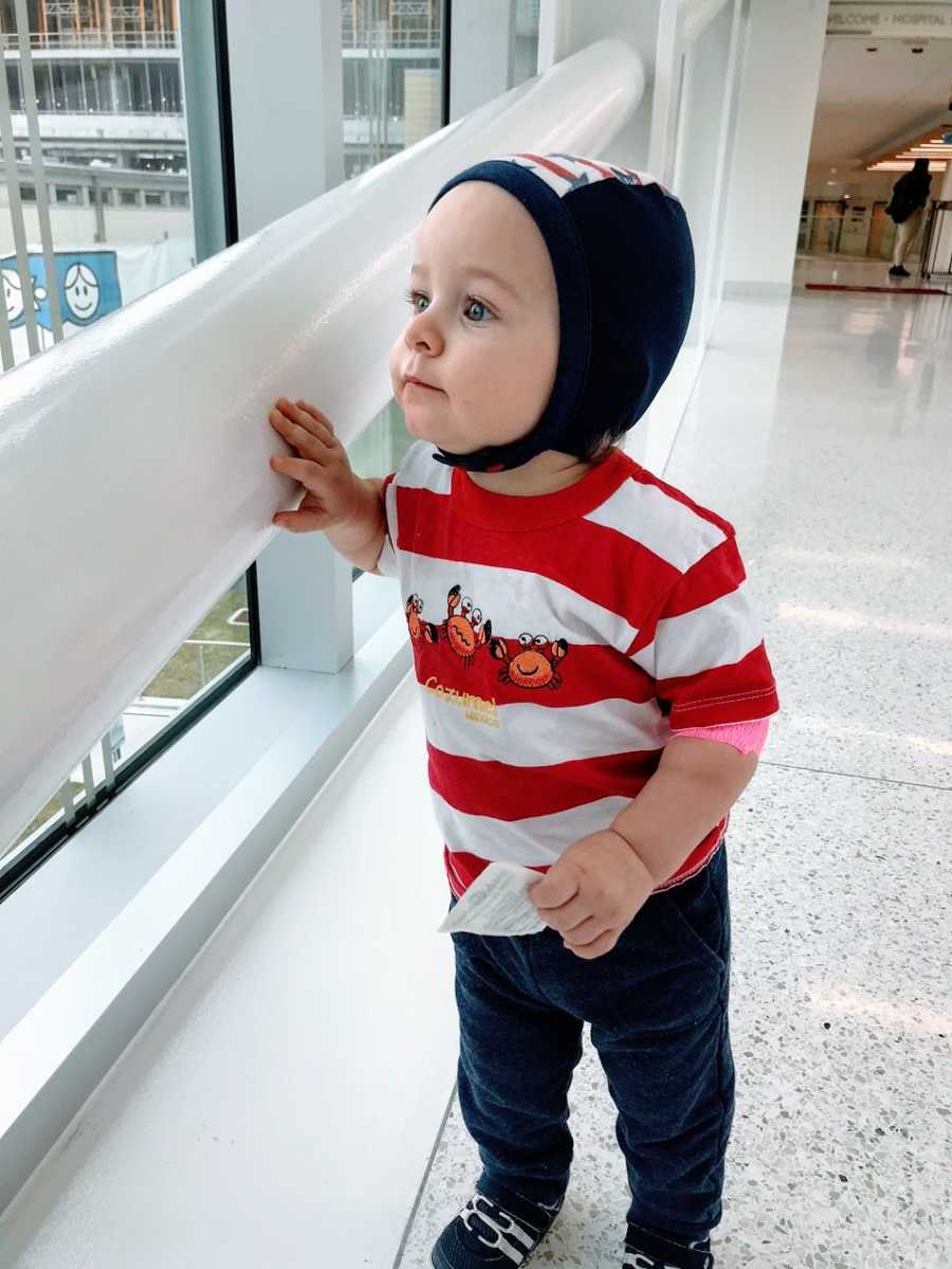 A little boy wearing a cap and a red striped shirt looks out a window