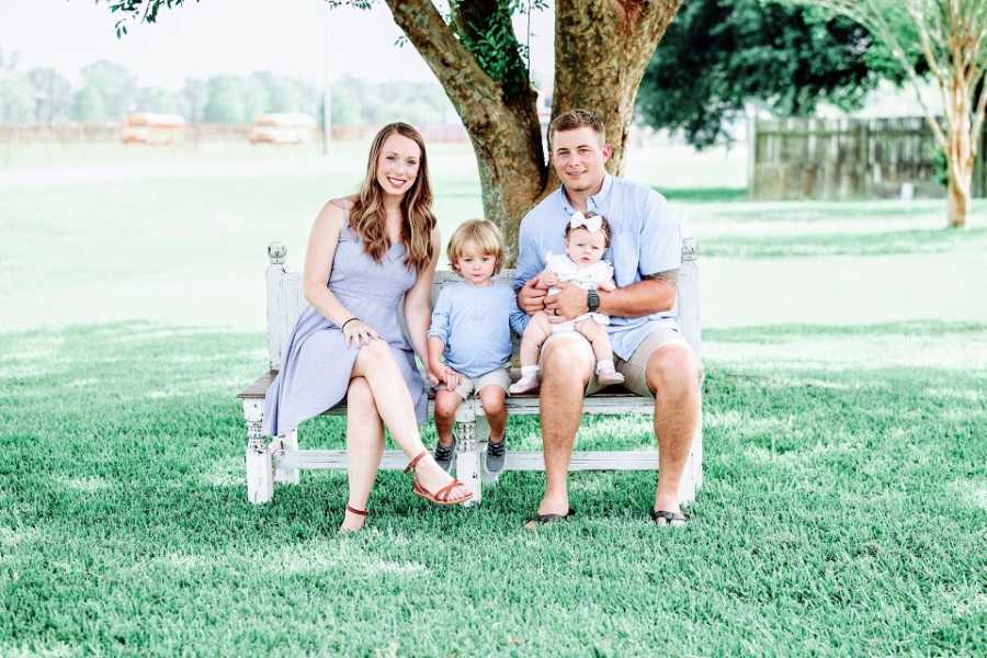 A couple sit on a park bench with their two young children