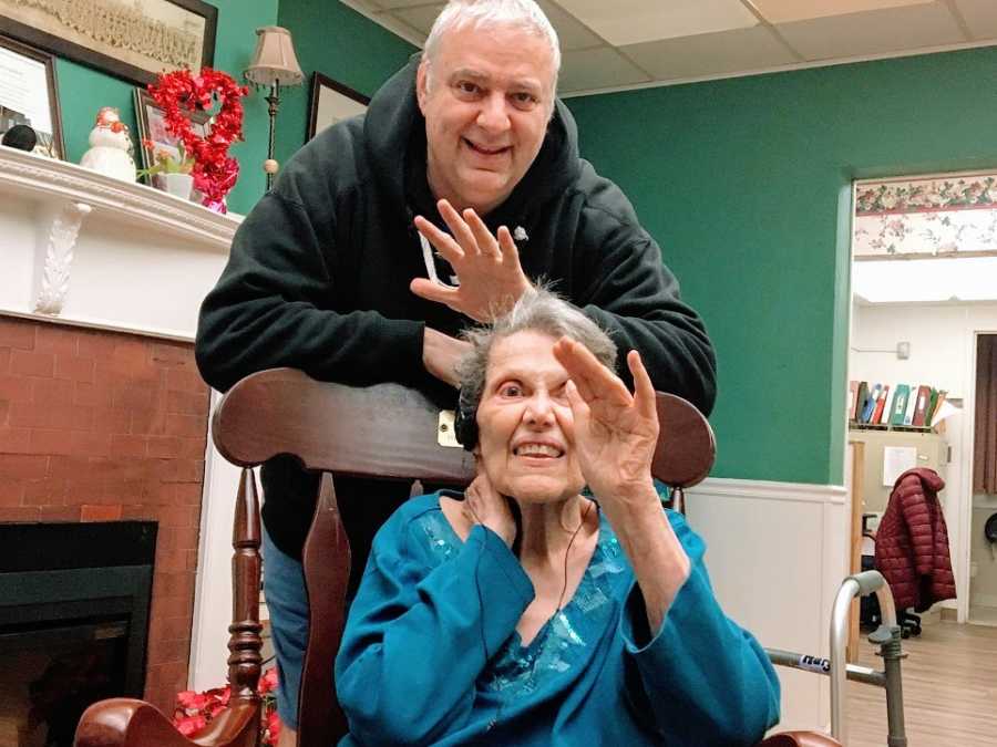 A man stands with an older woman who waves to the camera