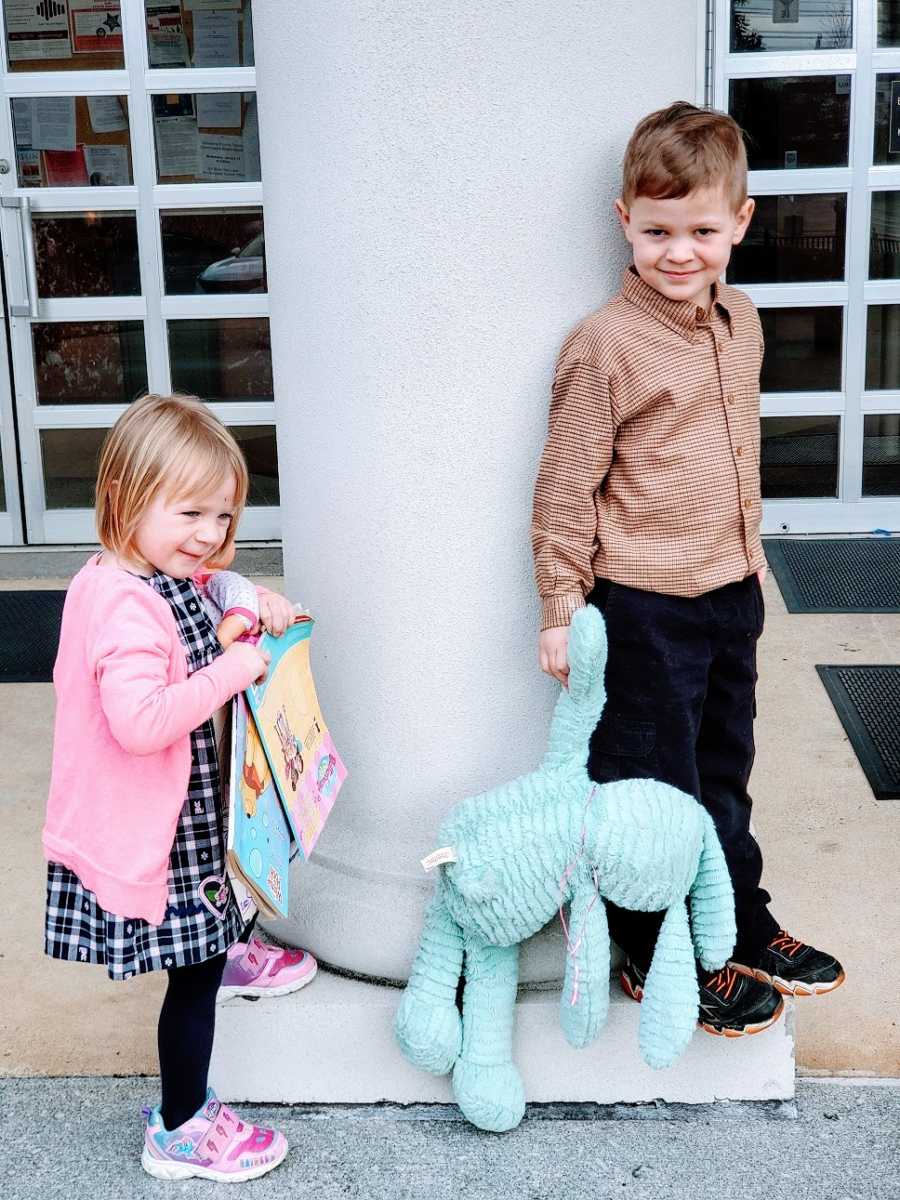 Siblings stand outside a building holding toys