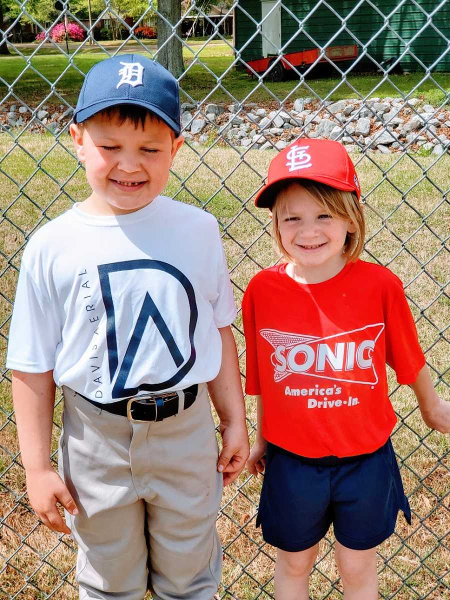 Siblings stand by a fence wearing baseball uniforms