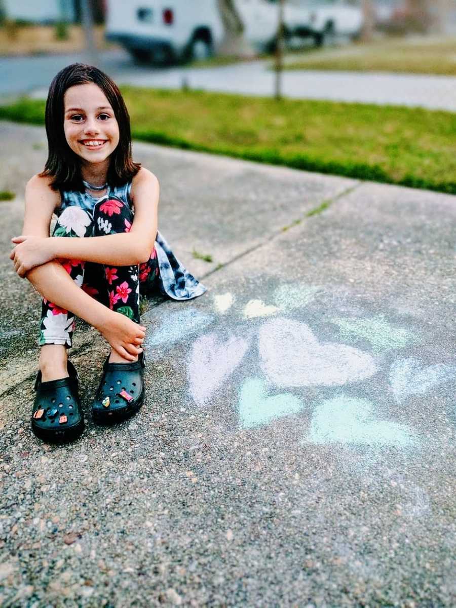 A little girl wearing crocs sits on a sidewalk next to chalk hearts