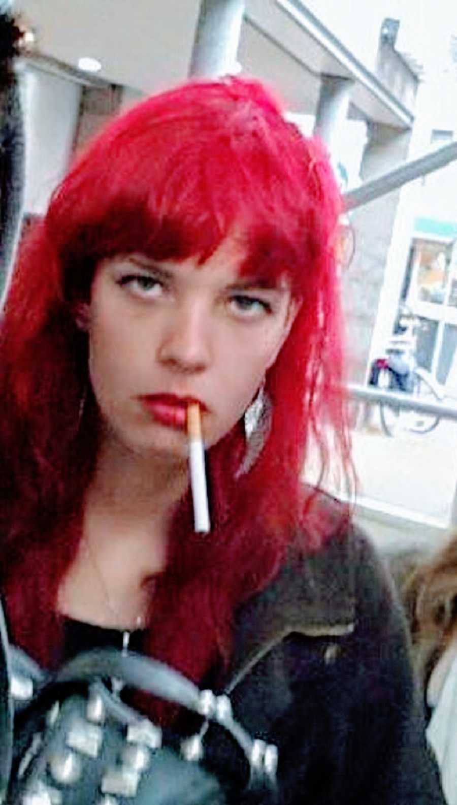 A young person with red hair holds a cigarette between their lips
