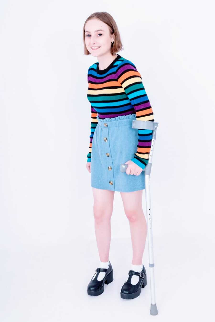 A young woman wearing a rainbow striped shirt stands with the help of a walking stick 