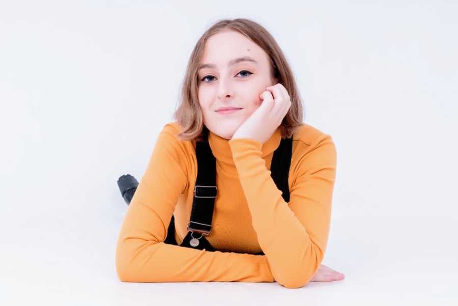 A young woman wearing a yellow shirt and overalls rests her head on her elbow