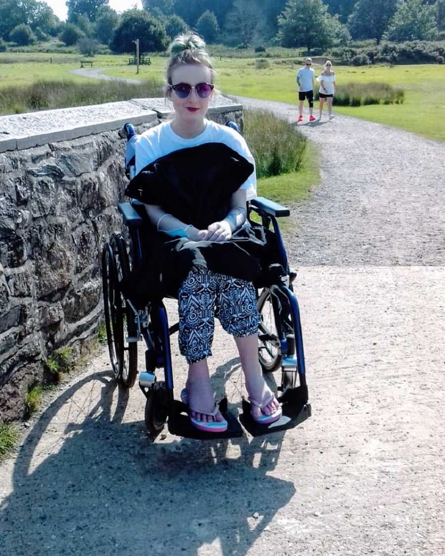 A woman sits in a wheelchair next to a stone wall wearing flipflops and sunglasses