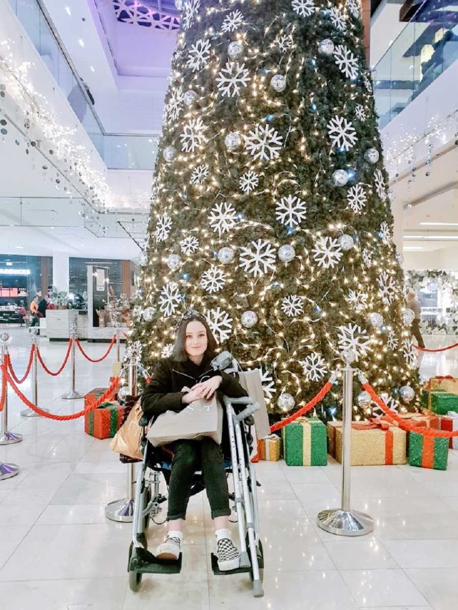 A woman in a wheelchair holds shopping bags near a large Christmas tree