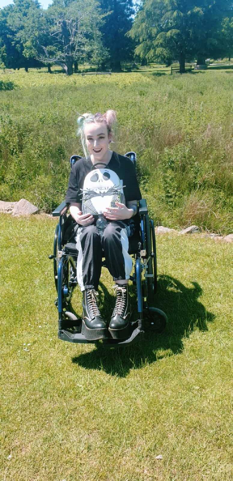 A woman in a wheelchair sits in a field holding a backpack