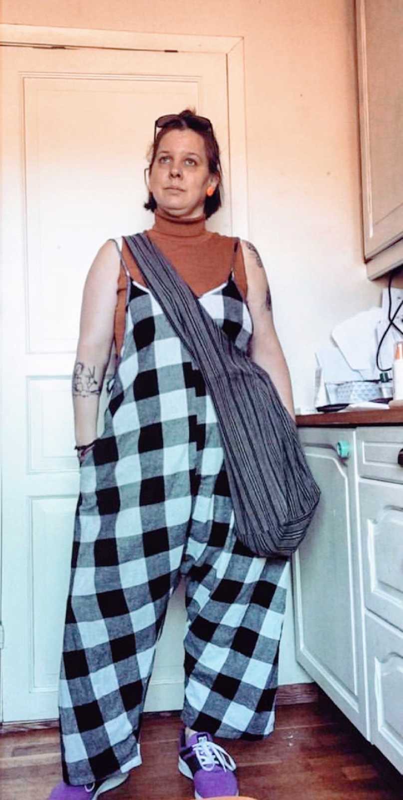 A nonbinary person wearing a pair of checkered overalls with hands in pockets