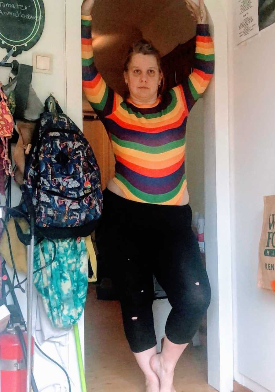 A nonbinary person wearing a rainbow striped shirt stands in a doorframe