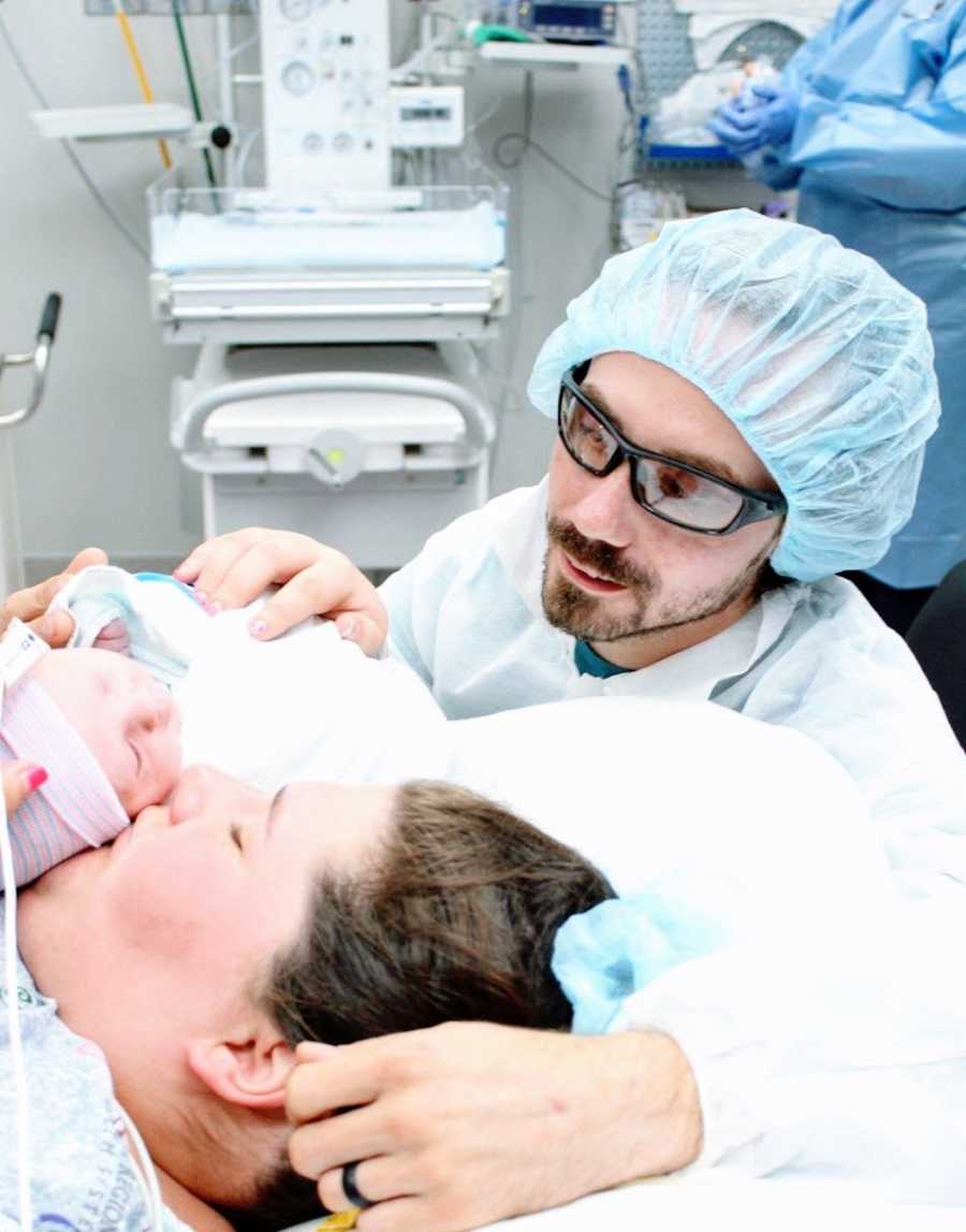 A husband wearing scrubs stands by his wife who kisses her newborn