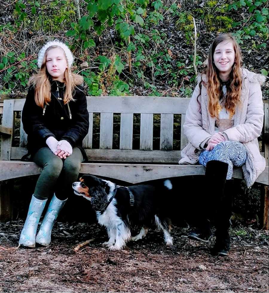 A pair of siblings sit on a park bench with a small dog between them