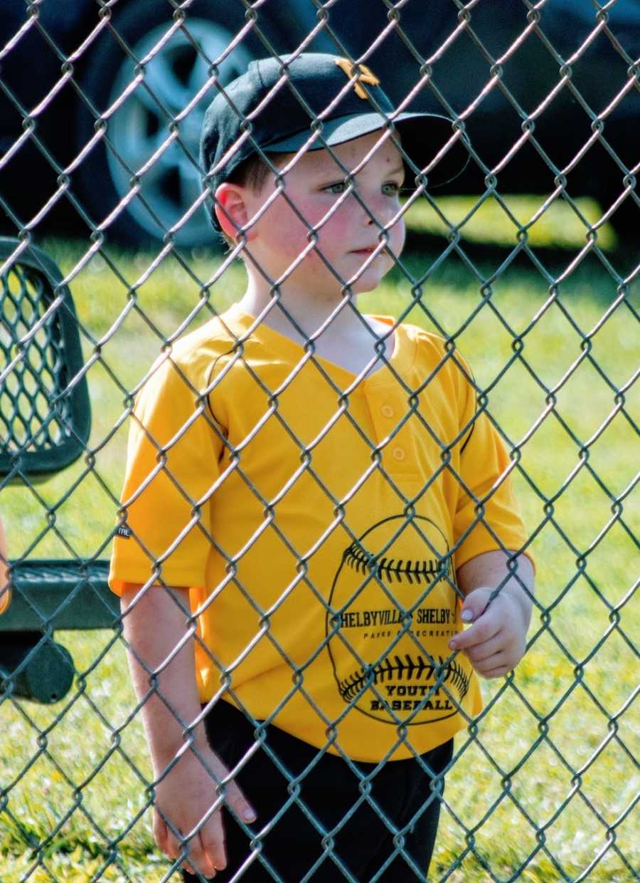 A boy wearing a baseball cap and yellow shirt stand behind a fence