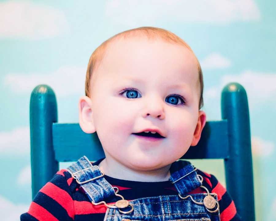 A baby boy wearing overalls and red stripes sits in a blue chair