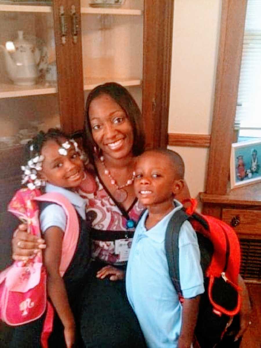 A mother stands with her two young children who wear backpacks