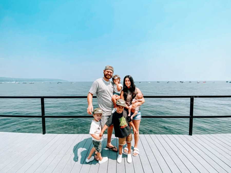 Couple take a family photo with their four sons under five while out on a walk on a boardwalk with the water behind them