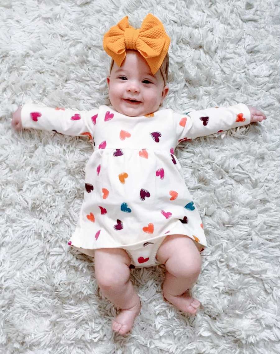 Young mom snaps photo of her miracle rainbow daughter smiling while laying on the floor in a heart onesie and a yellow bow on her head