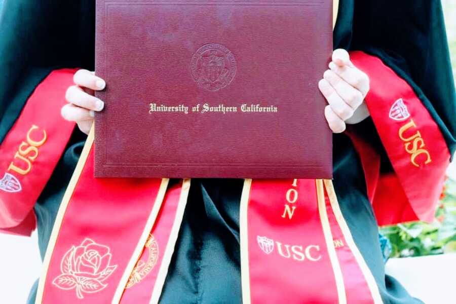 College student graduating from University of Southern California magna cum laude takes a photo with her diploma in her official robes and graduation gown