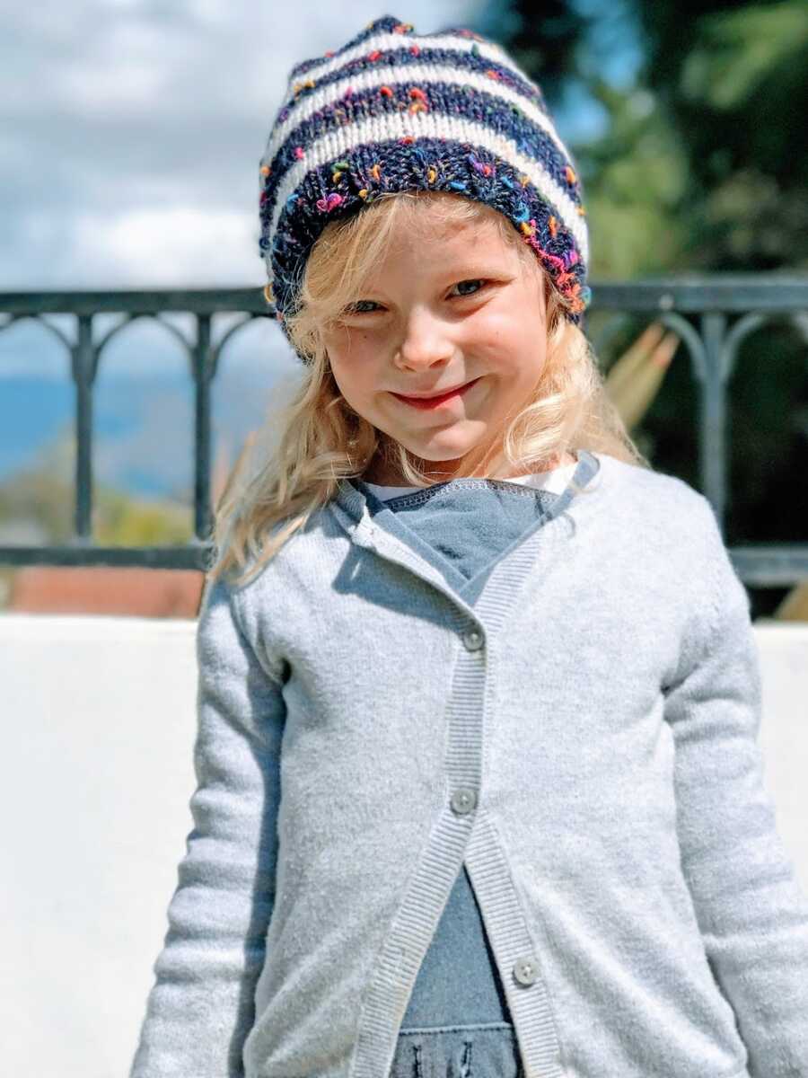 Little girl smiles big for a photo in a gray cardigan and speckled beanie
