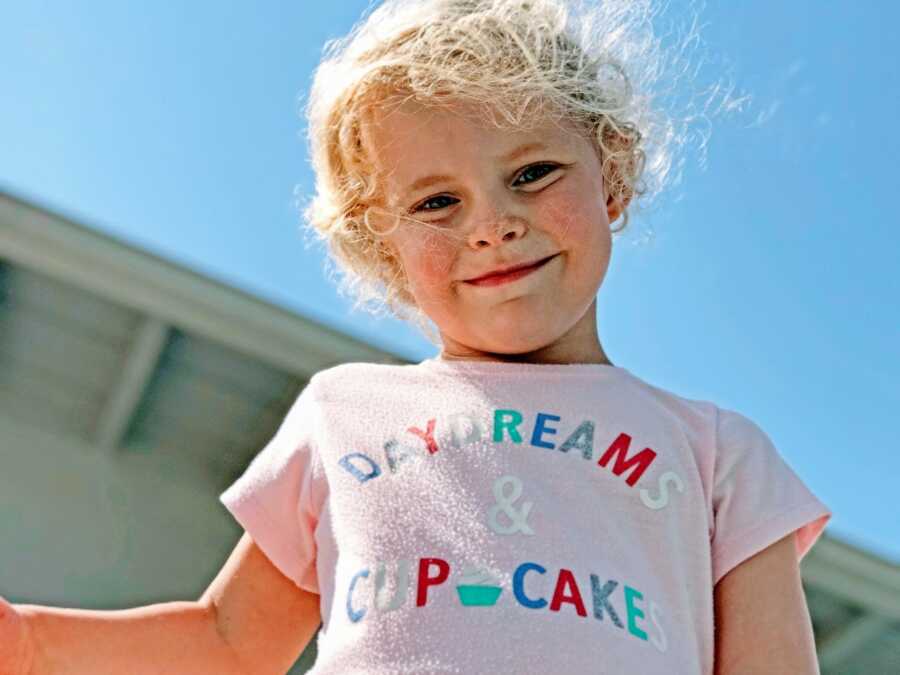 Little girl smiles down at a camera while wearing a pink shirt that says "Daydreams & Cupcakes"