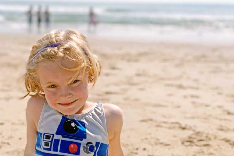 Young girl with a blonde bob smiles on the beach in an R2-D2 one-piece bathing suit