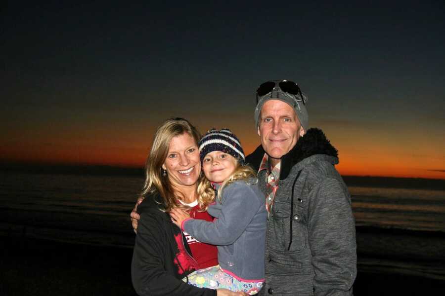 Parents smile big for a photo with their only daughter during the sunset on a beach while bundled up in winter clothes