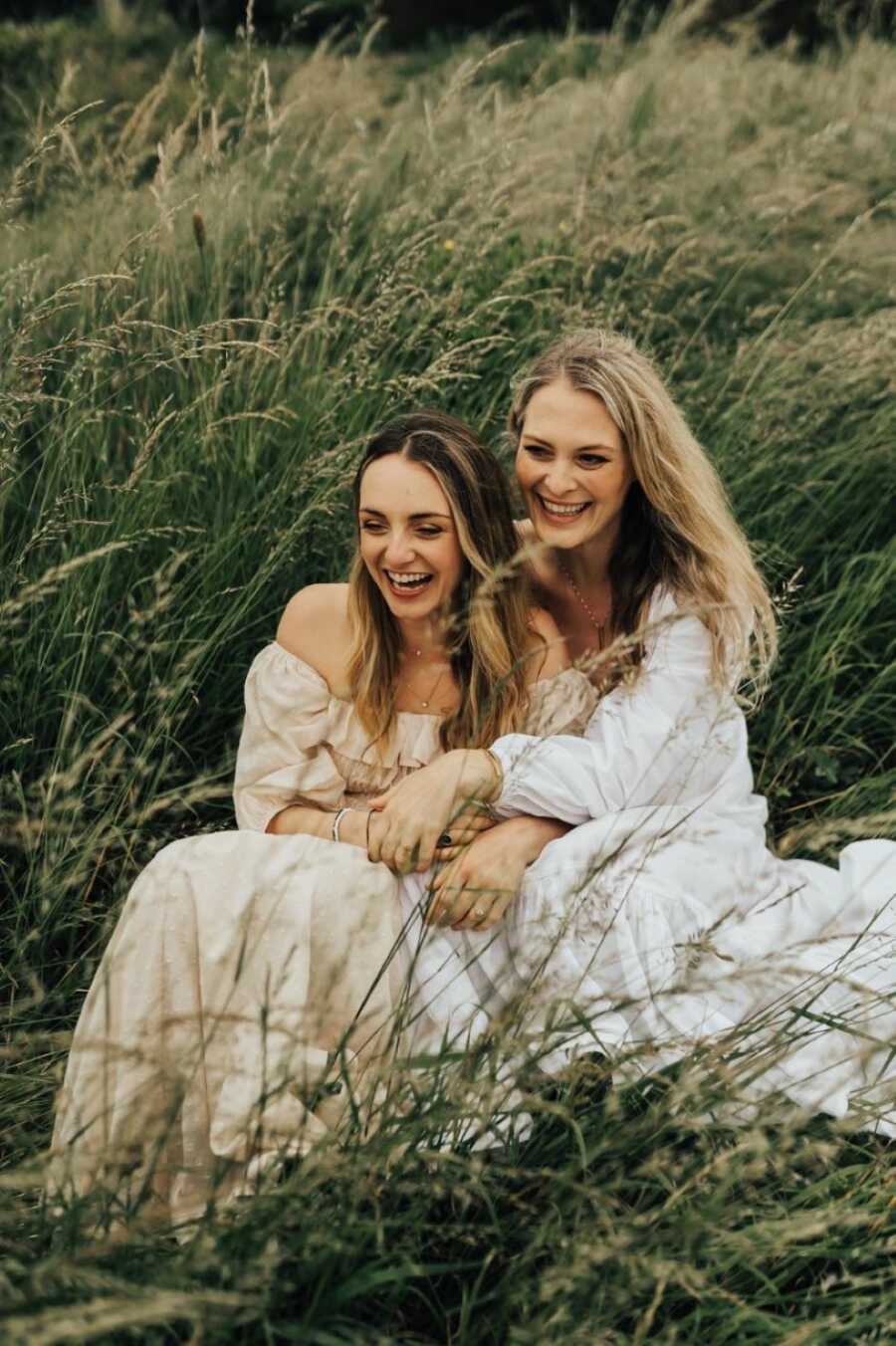 Two women engaged to be married wear long flowy dresses while taking photos in a field of wheat