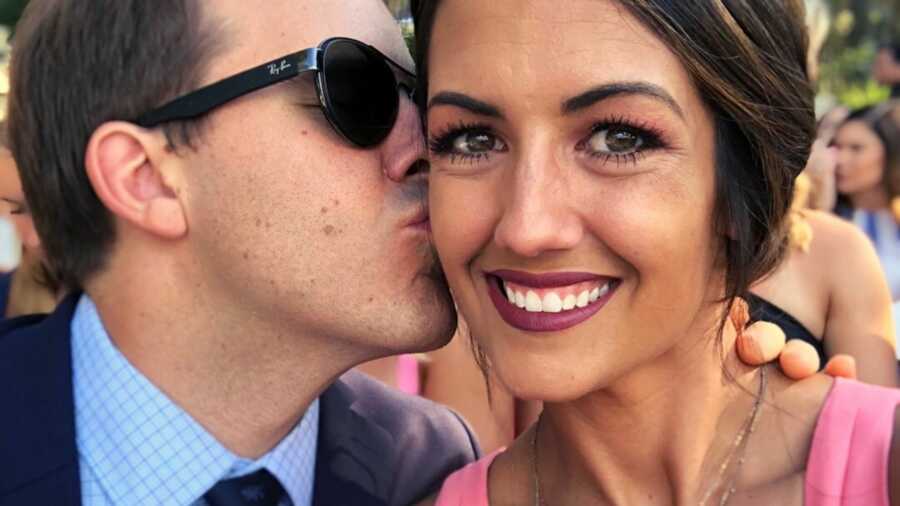 Man in sunglasses and tuxedo kissing cheek of woman in pink dress