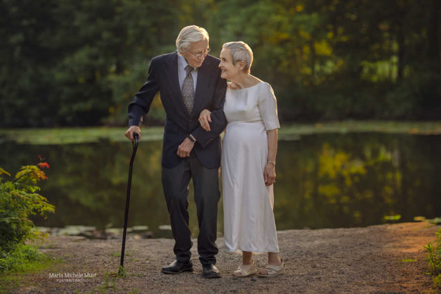 Elderly couple celebrating their 50th wedding anniversary walk arm-in-arm next to a lake, him in a blue suit and her in her original wedding dress