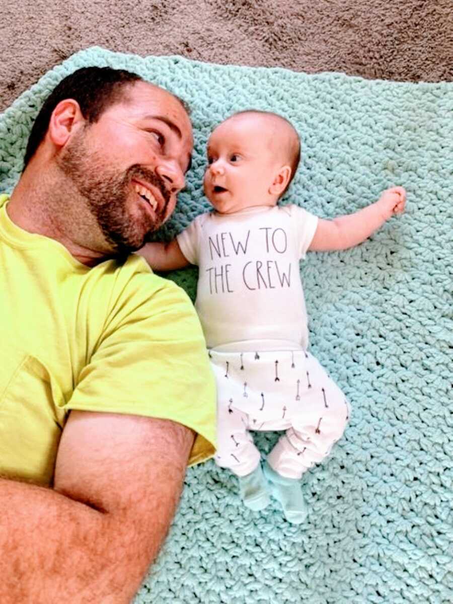 Dad lays on a blanket on the floor with his newborn daughter wearing a onesie that says "new to the crew"