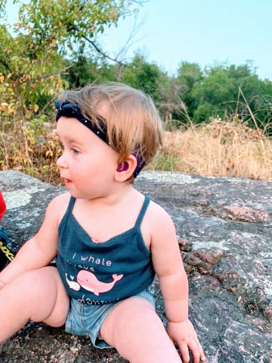 Little girl with hydrocephalus and deafness sits on a rock outside with hearing aids in