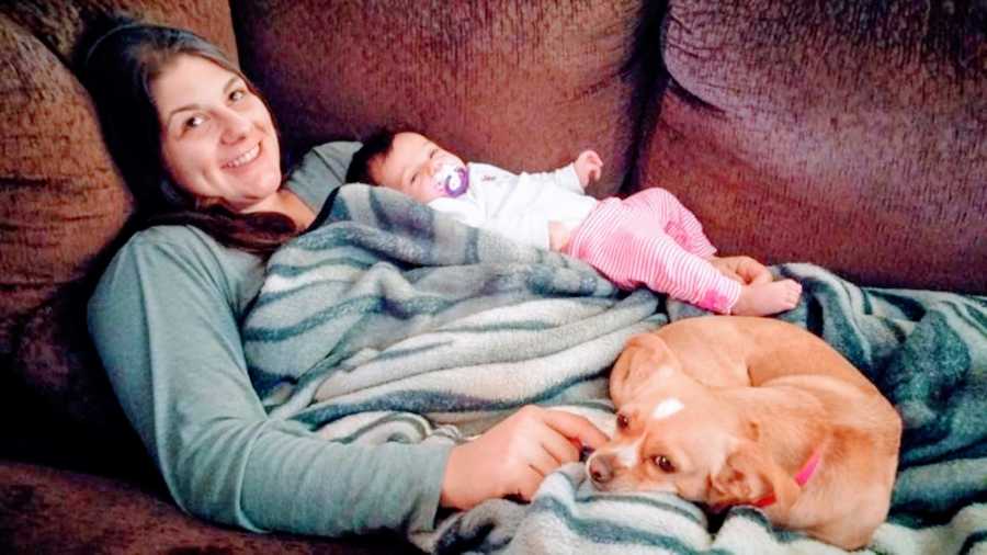 Young mom lays on a brown couch with her newborn daughter and her small brown dog