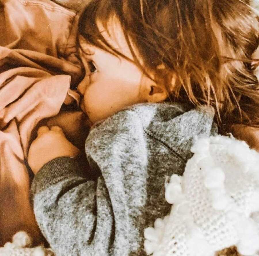 Little toddler with wavy brown hair cuddles up to their mom while breastfeeding