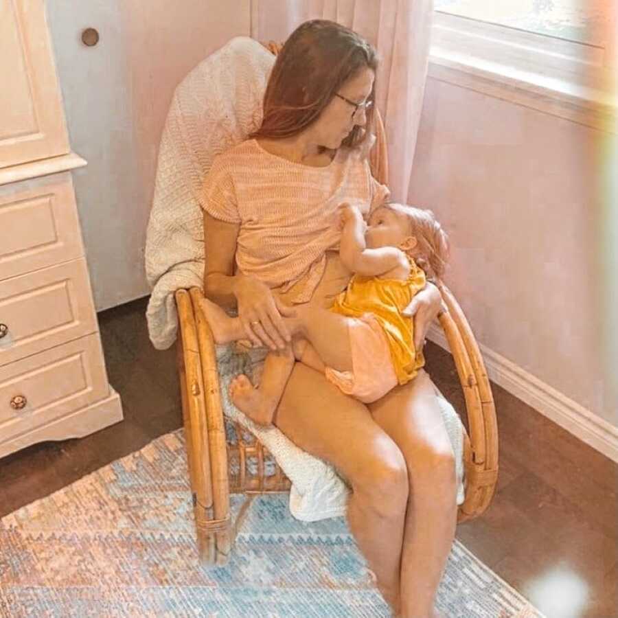 Mom sits in a wooden rocking chair while she breastfeeds her toddler