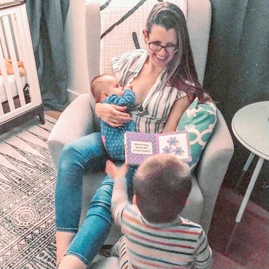 Mom breastfeeds her newborn in their nursery while reading her toddler a book