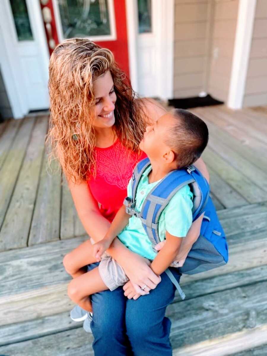 Mom and adopted son smile at each other while he sits on her lap with a backpack on before his first day of school