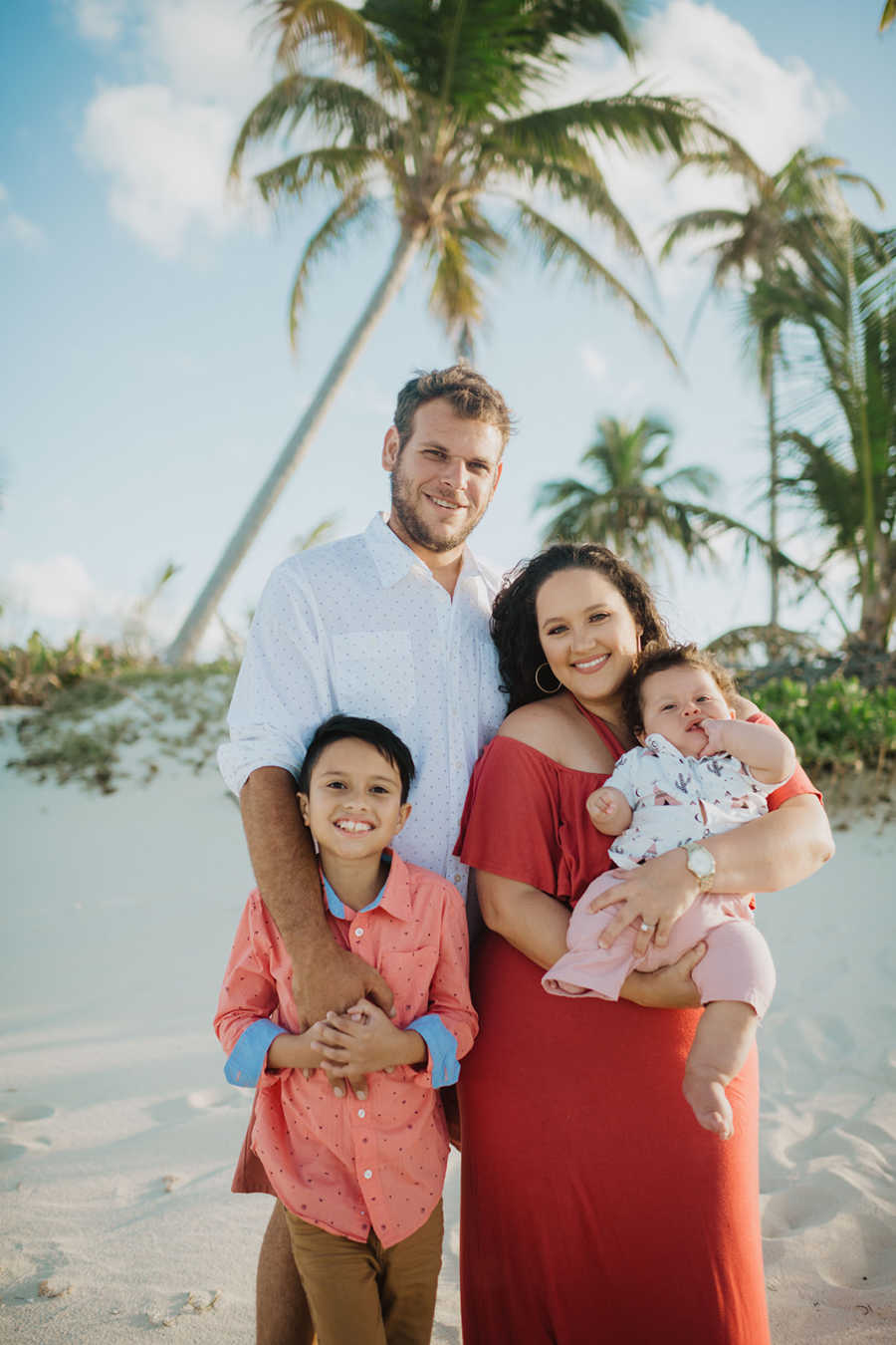 Family of four living in the Bahamas take a family photo together on a white sandy beach with palm trees behind them