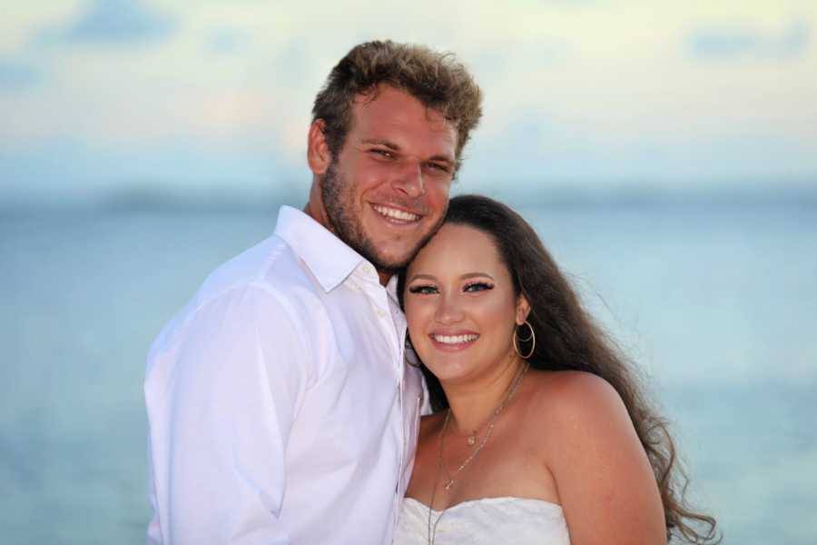 Young couple take maternity photos while wearing white on the beach at sunset