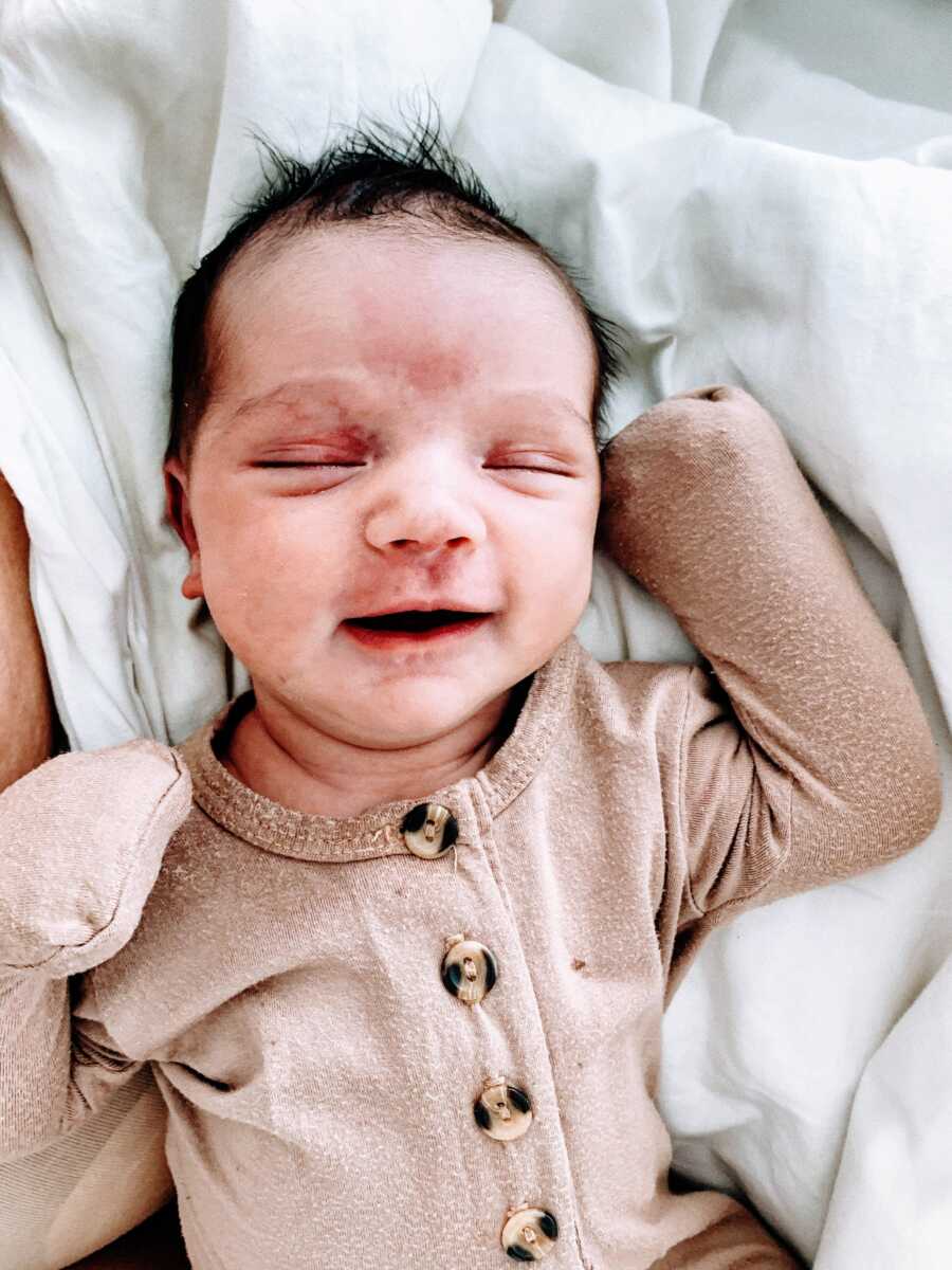 Newborn baby boy with a head full of dark hair in tan onesie smiles big in his sleep