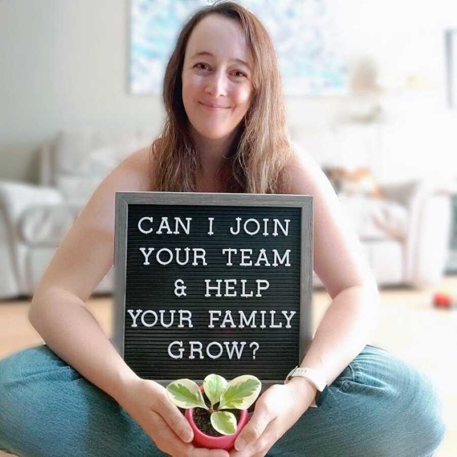 Mom of 2 looking to become a surrogate takes a photo with a plant and a sign that reads "Can I join your team & help your family grow?"