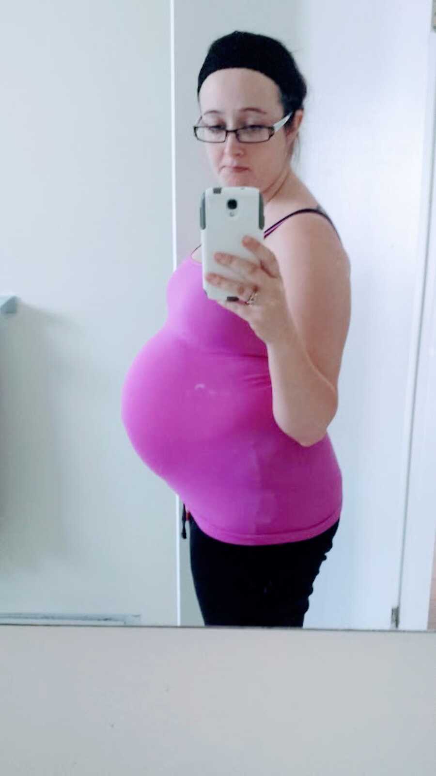 Heavily pregnant woman takes a mirror selfie, showing off her belly bump in a purple tanktop