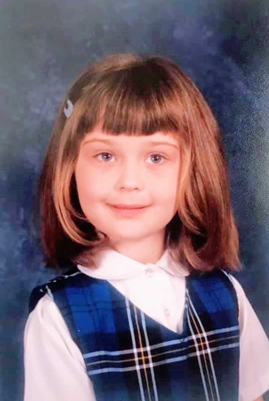Little girl who with straight across bangs smiles in a school uniform for her school yearbook photo