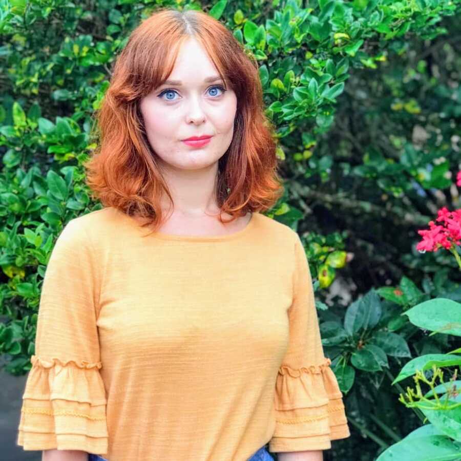 Young woman discovering her autistic and ADHD identities poses for a photo in front of red flowers in a yellow shirt