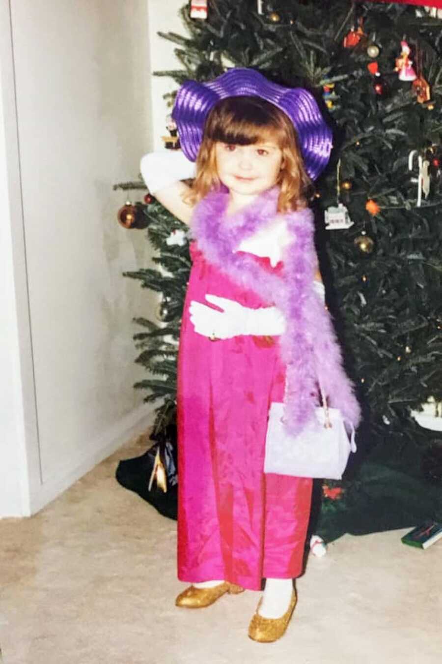 Little girl dressed in a pink dress with a purple hat, feather boa, purse, and white gloves poses in front of a Christmas tree