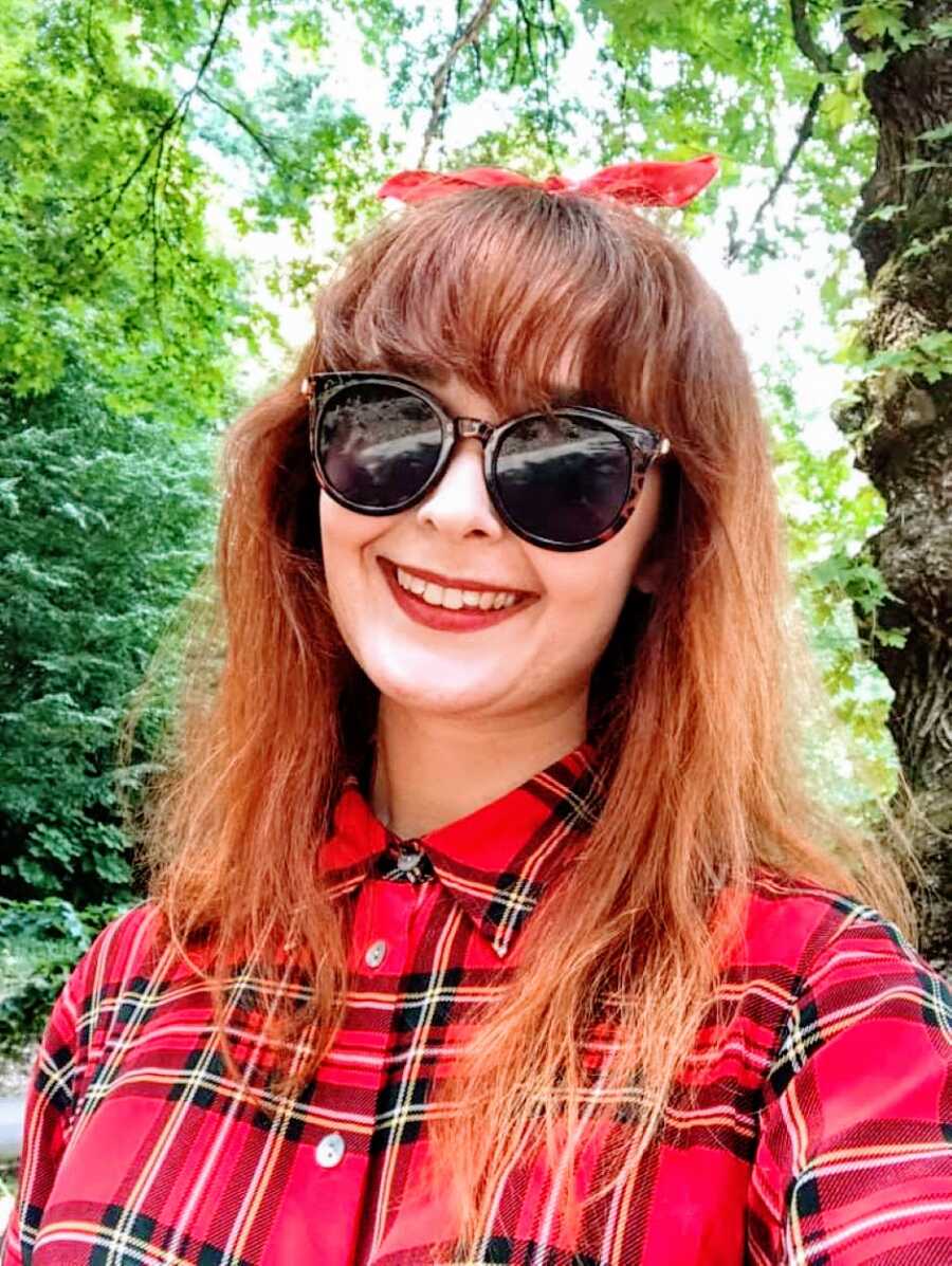 Young woman with autism and ADHD takes a selfie in a red plaid shirt with sunglasses on and a red bow in her hair