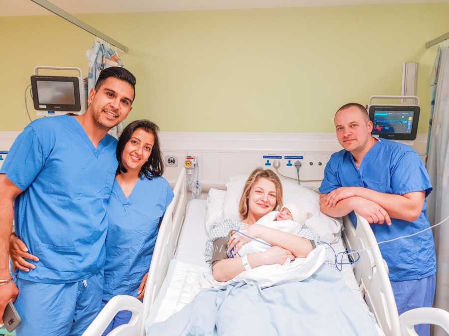 Surrogate holds newborn baby she just birthed while the intended parents smile next to her in scrubs